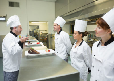 Three chefs presenting their dessert plates to the head chef in busy kitchen